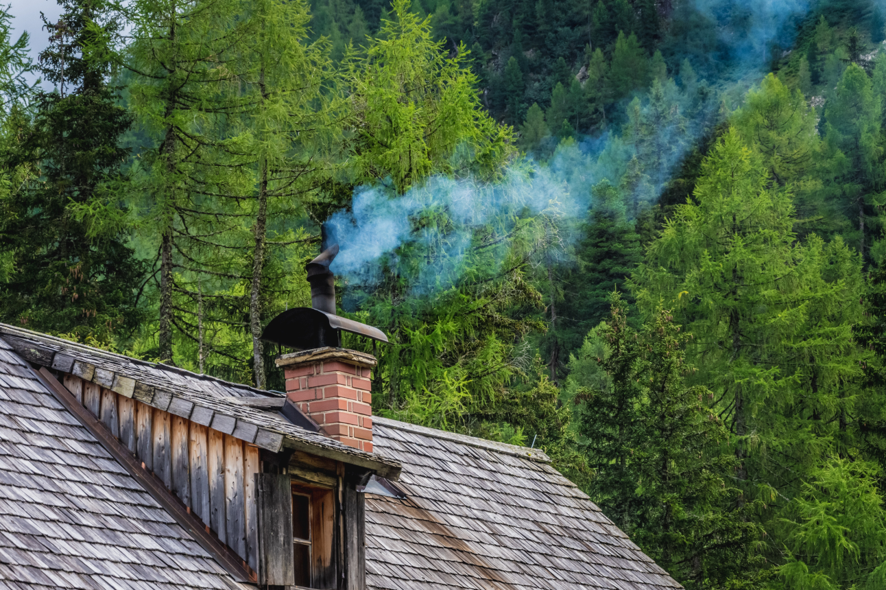 Bild von einem Schornstein, einem Hausdach und im Hintergrund ist ein wunderschöner grüner Tannenwald. Die Ziegel des Dachs sind rötlich braun.