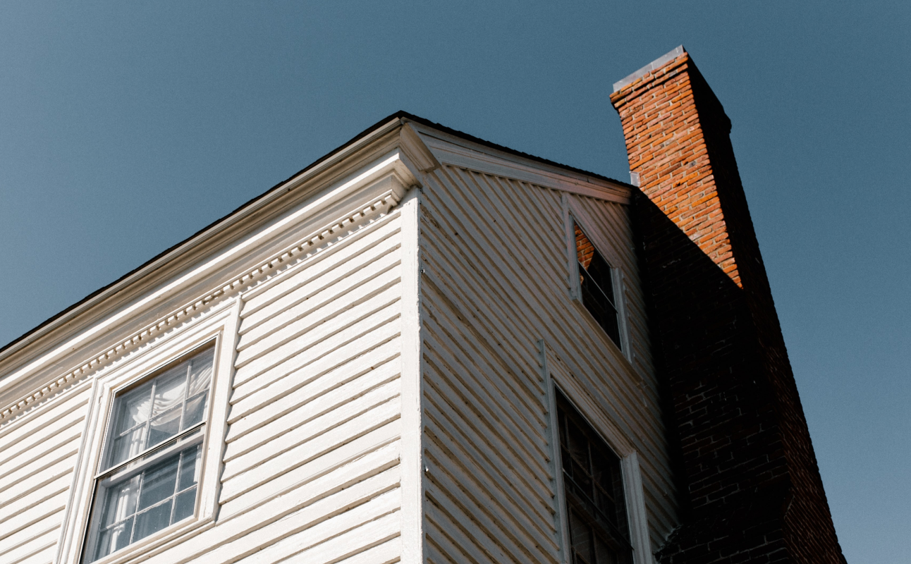 Ein weißes Haus, mit zwei sichtbaren Fenstern und einem roten Schornstein. Von schräg unten nach oben fotografiert. Der Himmel ist grau blau